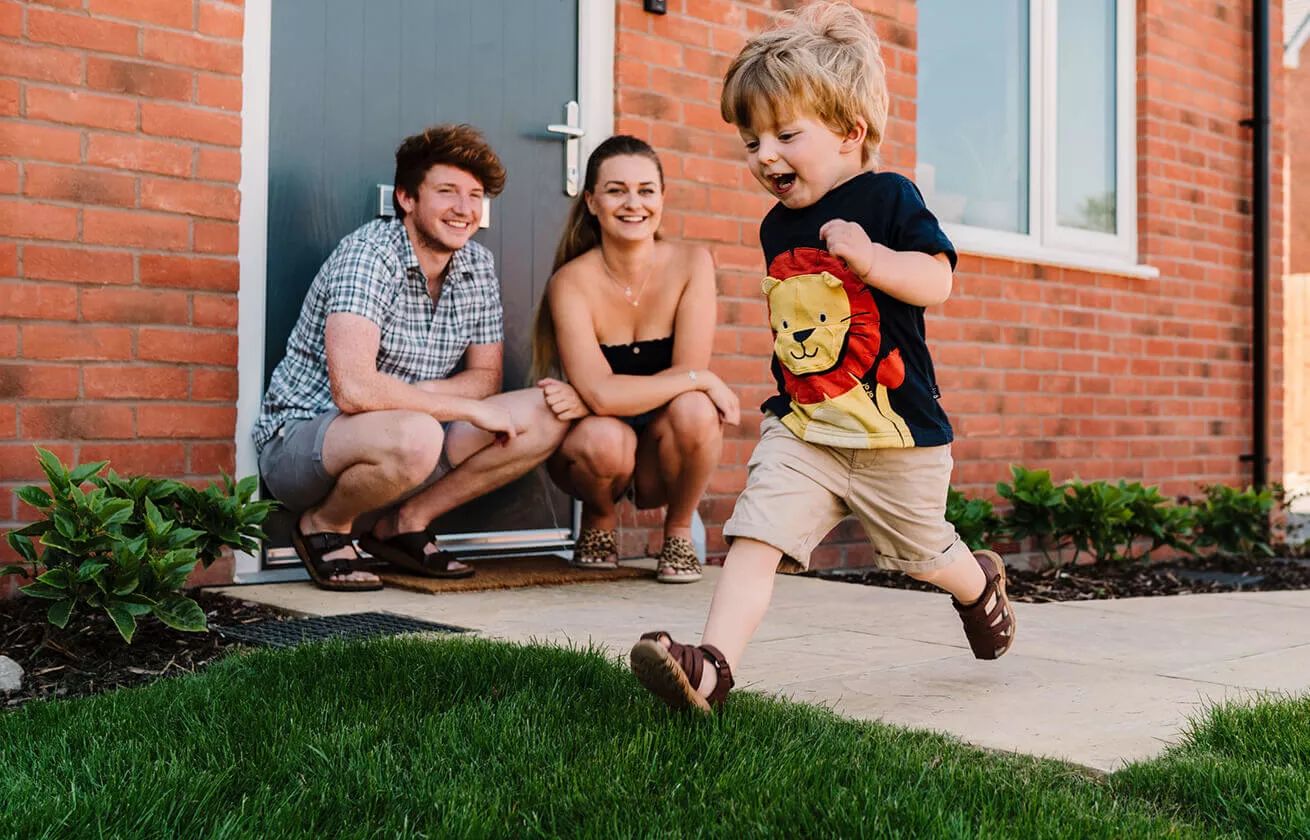 A young family outside a new house, the 2 parents are squatting down whilst a toddler runs